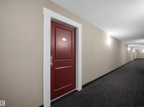 Hallway featuring a paneled red entry door with chrome hardware, white trim, light beige walls, and dark striped carpeting - #422 5515 7 Av Sw, Edmonton, AB - Indoor Photo Showing Other Room