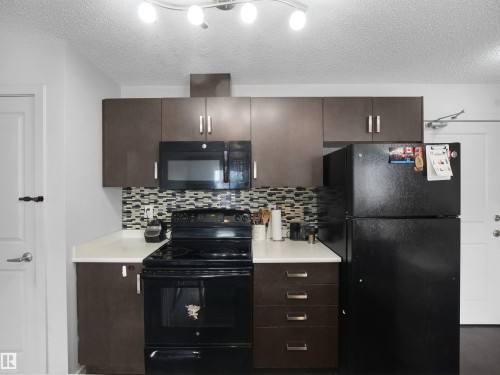 Contemporary kitchen featuring dark wood-finish cabinetry, white countertops, a mosaic tile backsplash, and black appliances including a microwave and range - #422 5515 7 Av Sw, Edmonton, AB - Indoor Photo Showing Kitchen