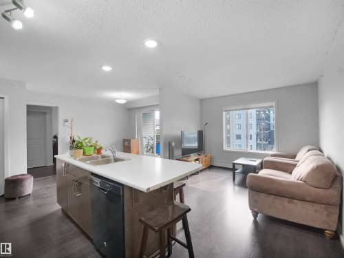 Open-concept living space featuring dark wood-finish flooring, recessed lighting, a kitchen island with a double basin sink, and a sliding glass door to a balcony - #422 5515 7 Av Sw, Edmonton, AB - Indoor