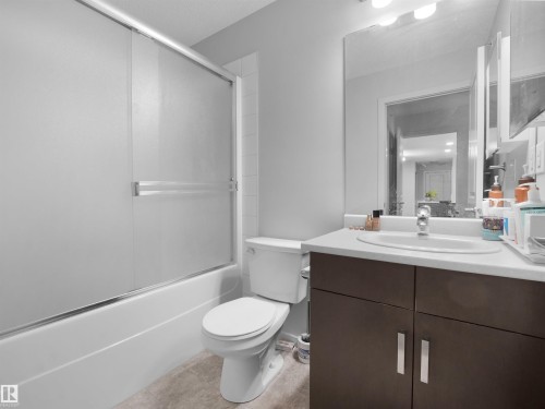 Bathroom featuring a tub and shower combination with frosted glass sliding doors, a single vanity with a white countertop and integrated sink, a large wall-mounted mirror, and dark wood-finish cabinetry with silver hardware - #422 5515 7 Av Sw, Edmonton, AB - Indoor Photo Showing Bathroom