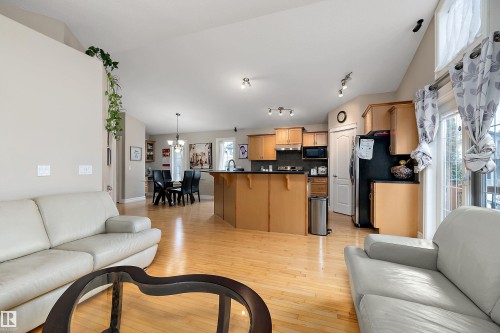 Open-concept living space with light wood-finish flooring, a kitchen featuring wood cabinetry and black appliances, a dining area with a chandelier, vaulted ceilings, and a sliding glass door to exterior - 16748 118 Street Nw, Edmonton, AB - Indoor Photo Showing Living Room