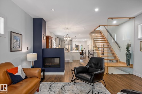Open-concept living space featuring wood-finish flooring, a contemporary fireplace with a navy surround, and a modern staircase with glass railing - 14221 101 Avenue Nw, Edmonton, AB - Indoor Photo Showing Living Room With Fireplace