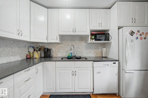 Kitchen featuring white cabinetry with silver hardware, dark countertops, a mosaic tile backsplash, stainless steel sink with a gooseneck faucet, and wood-finish flooring - 18407 95A Avenue Nw, Edmonton, AB - Indoor Photo Showing Kitchen