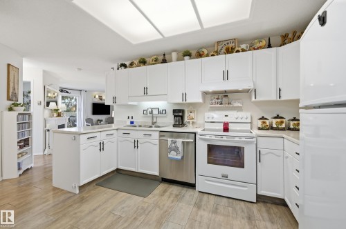 Bright kitchen featuring white cabinetry with black hardware, stainless steel dishwasher, white range, and wood-finish flooring - #208 9760 176 St Nw, Edmonton, AB - Indoor Photo Showing Kitchen