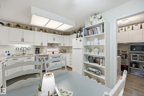 White kitchen cabinetry with dark hardware, a surface-mounted ceiling light fixture, wood-finish flooring, and a built-in bookshelf - #208 9760 176 St Nw, Edmonton, AB - Indoor Photo Showing Kitchen With Double Sink