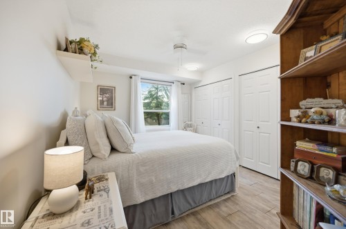 Bedroom featuring wood-finish flooring, a window with exterior tree canopy, two bi-fold closet doors, a ceiling fan, and a built-in wall shelf - #208 9760 176 St Nw, Edmonton, AB - Indoor Photo Showing Bedroom