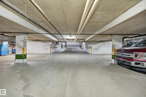 Underground parking garage featuring concrete flooring, exposed overhead piping, structural columns with protective markings, and overhead lighting - #208 9760 176 St Nw, Edmonton, AB - Indoor Photo Showing Garage