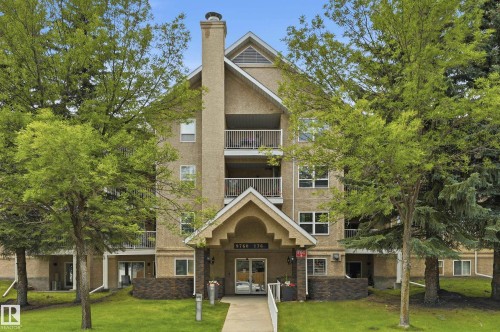 Multi-story residential building featuring a stucco facade, multiple balconies, a covered entryway with stone-finish accents, and mature trees framing the structure - #208 9760 176 St Nw, Edmonton, AB - Outdoor With Facade