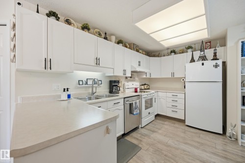 Contemporary kitchen featuring white cabinetry with black hardware, a dual-basin stainless steel sink, and light-toned countertops - #208 9760 176 St Nw, Edmonton, AB - Indoor Photo Showing Kitchen With Double Sink
