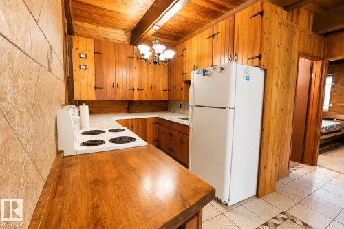 Kitchen featuring wood-paneled walls and ceiling, exposed wooden beams, U-shaped light-toned countertops, wood-finish cabinetry with decorative black hinges, and tile flooring - 329 60417 Range Road 124, Rural St. Paul County, AB - Indoor Photo Showing Kitchen