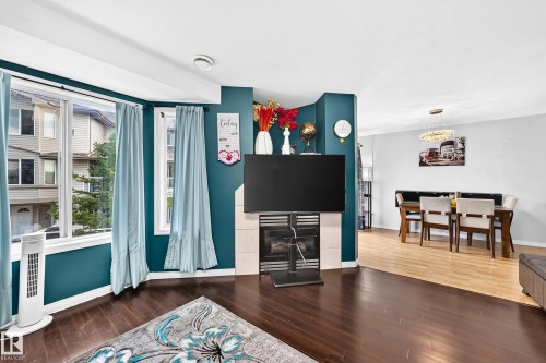 Living area featuring a fireplace with a light-toned surround, dark wood-finish flooring, and a large bay window - #72 2505 42 St Nw, Edmonton, AB - Indoor Photo Showing Living Room With Fireplace