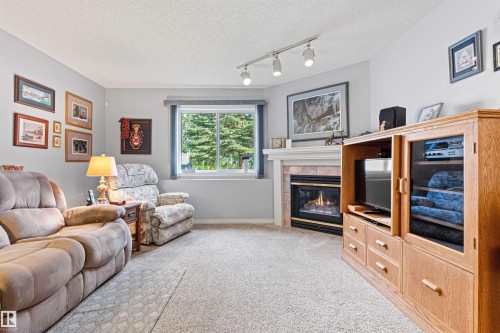 Family room featuring a gas fireplace with a tile surround and white mantelpiece, track lighting, a window, and light-toned carpeting - #12 64 Blackburn Dr W Sw, Edmonton, AB - Indoor Photo Showing Living Room With Fireplace
