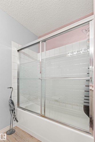 Bathtub with sliding glass enclosure featuring frosted privacy panels, white subway tile surround, and a textured ceiling - #12 64 Blackburn Dr W Sw, Edmonton, AB - Indoor Photo Showing Bathroom