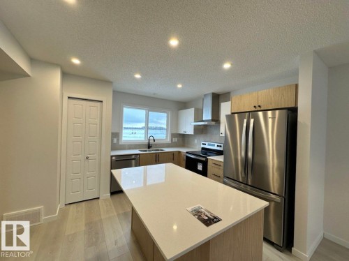 Modern kitchen featuring a large central island with a light-toned countertop, stainless steel appliances, wood-finish cabinetry, recessed ceiling lighting, and wood-finish flooring - 22803 94 Anenue Northwest, Edmonton, AB - Indoor Photo Showing Kitchen With Double Sink