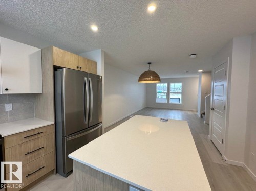 Open-concept kitchen featuring an oversized island with a light-toned countertop, stainless steel refrigerator, two-tone cabinetry with wood-finish and white doors, recessed lighting, and a woven pendant light fixture - 22803 94 Anenue Northwest, Edmonton, AB - Indoor Photo Showing Other Room