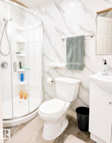 Bathroom featuring a curved glass shower enclosure with built-in shelving, white vanity with integrated sink, white toilet, and marbled wall panels - 3236 27 Avenue Nw, Edmonton, AB - Indoor Photo Showing Bathroom