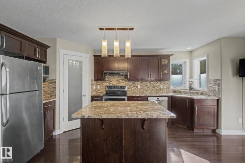 Kitchen featuring dark wood cabinetry, a granite-topped island, stainless steel appliances, a mosaic tile backsplash, and wood-finish flooring - 1047 Leger Boulevard Nw, Edmonton, AB - Indoor Photo Showing Kitchen With Stainless Steel Kitchen With Double Sink With Upgraded Kitchen