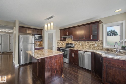 Kitchen featuring dark wood cabinetry, stainless steel appliances, a mosaic tile backsplash, wood-finish flooring, and a central island with a light-toned countertop - 1047 Leger Boulevard Nw, Edmonton, AB - Indoor Photo Showing Kitchen With Stainless Steel Kitchen With Double Sink With Upgraded Kitchen