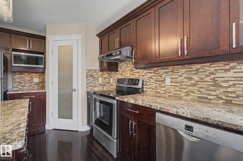Kitchen featuring dark wood cabinetry, light-toned granite-style countertops, stainless steel appliances, a mosaic tile backsplash, and dark wood-finish flooring - 1047 Leger Boulevard Nw, Edmonton, AB - Indoor Photo Showing Kitchen With Stainless Steel Kitchen With Upgraded Kitchen