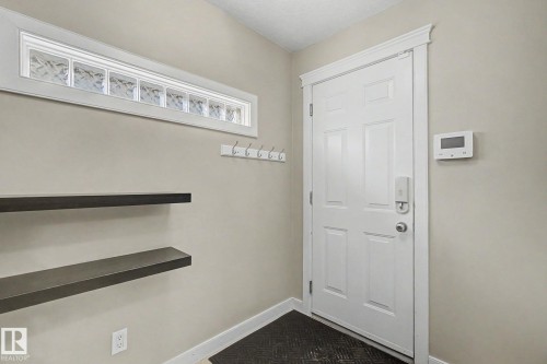 Entryway featuring a white paneled door, a transom window with patterned glass blocks, two dark-toned floating shelves, a row of white coat hooks, and a wall-mounted control panel - 1047 Leger Boulevard Nw, Edmonton, AB - Indoor Photo Showing Other Room