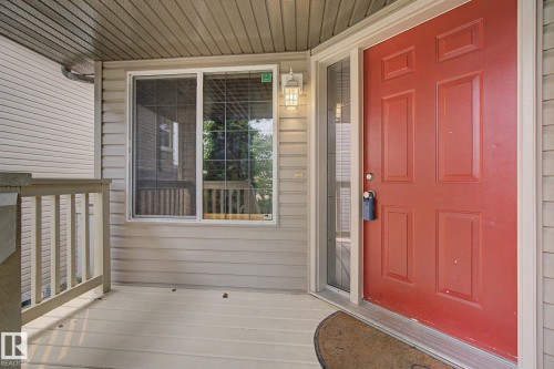 Front entry featuring a red paneled door with a sidelight, covered porch, light beige siding, and a white outdoor wall sconce - 1047 Leger Boulevard Nw, Edmonton, AB - Outdoor With Deck Patio Veranda With Exterior