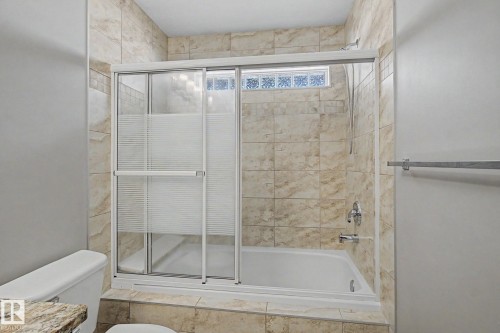 Bathtub and shower combination featuring a glass slider enclosure with frosted panels, light-toned stone tile surround, and a chrome fixture - 1047 Leger Boulevard Nw, Edmonton, AB - Indoor Photo Showing Bathroom
