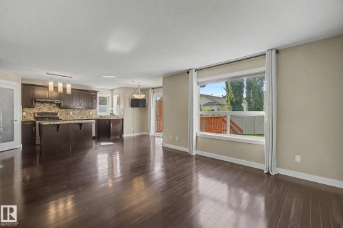 Spacious open-concept room featuring rich wood-finish flooring, a large picture window, and a kitchen with dark cabinetry, stone countertops, and a tile backsplash - 1047 Leger Boulevard Nw, Edmonton, AB - Indoor