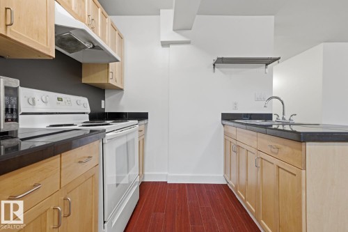Kitchen featuring wood-finish flooring, light wood cabinetry with brushed nickel hardware, dark countertops, and a stainless steel sink with a gooseneck faucet - #3 9955 114 St Nw, Edmonton, AB - Indoor Photo Showing Kitchen