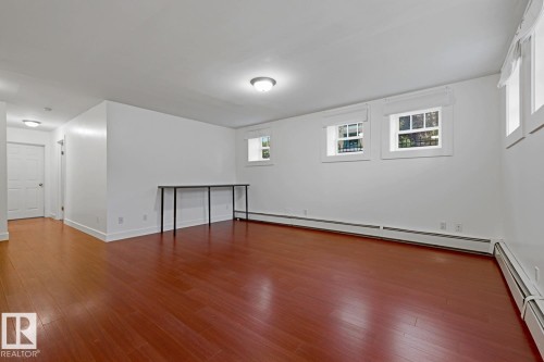 Spacious room featuring wood-finish flooring, white walls, multiple windows with white trim, and baseboard heating - #3 9955 114 St Nw, Edmonton, AB - Indoor Photo Showing Other Room