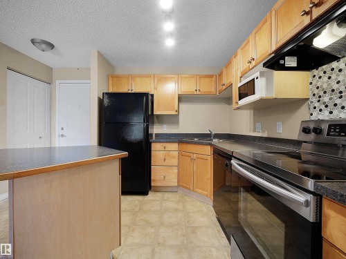 Kitchen featuring light wood cabinetry, dark countertops, a black appliance suite, a built-in microwave, and a decorative tile backsplash - #220 245 Edwards Dr Sw, Edmonton, AB - Indoor Photo Showing Kitchen
