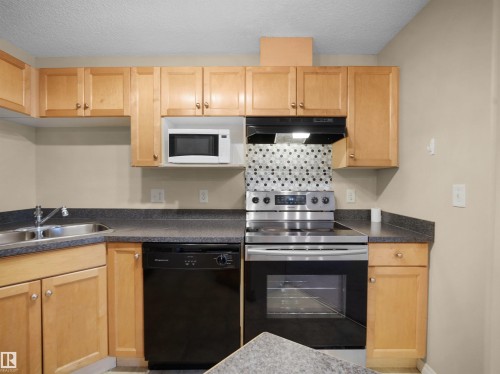 Kitchen featuring light wood cabinetry, dark speckled countertops, a stainless steel range, and a patterned tile backsplash - #220 245 Edwards Dr Sw, Edmonton, AB - Indoor Photo Showing Kitchen With Double Sink