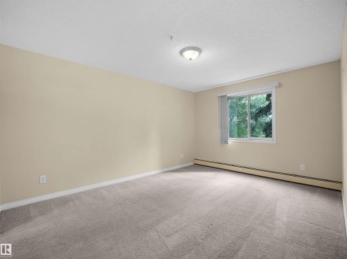 Spacious room featuring neutral carpeting, light-toned walls, and a large window with vertical blinds - #220 245 Edwards Dr Sw, Edmonton, AB - Indoor Photo Showing Other Room