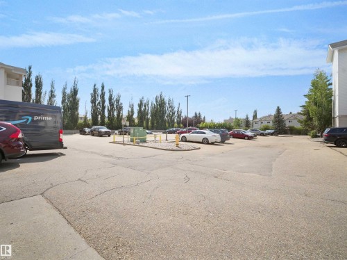Paved parking area featuring multiple vehicle spaces, mature trees along the perimeter, and adjacent residential structures - #220 245 Edwards Dr Sw, Edmonton, AB - Outdoor