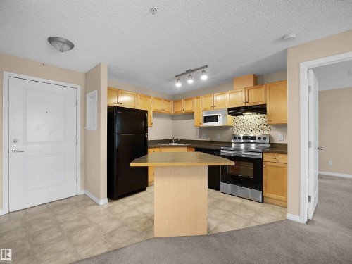Kitchen featuring wood cabinetry, stone-finish countertops, stainless steel appliances, and a tiled backsplash - #220 245 Edwards Dr Sw, Edmonton, AB - Indoor Photo Showing Kitchen