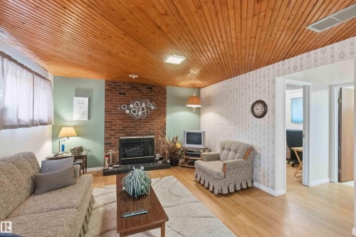 Wood-finish plank ceiling with integrated lighting, brick fireplace with a black hearth, light wood-finish flooring, and a window with sheer coverings - 30 Cavanagh Crescent, Stony Plain, AB - Indoor Photo Showing Living Room With Fireplace