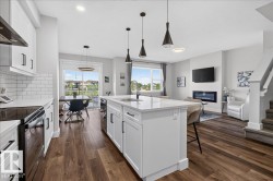Open-concept living space featuring a central kitchen island with a built-in sink, wood-finish flooring, white cabinetry with black hardware, a tiled backsplash, and an electric fireplace - 