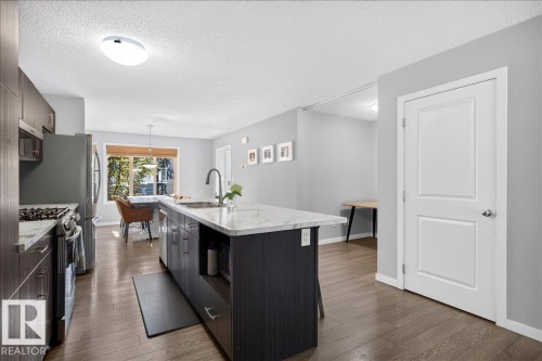 Contemporary kitchen featuring a large central island with a stone-finish countertop, dark wood-finish cabinetry, stainless steel appliances, and wood-finish flooring - 3043 Keswick Way Sw, Edmonton, AB - Indoor Photo Showing Kitchen With Upgraded Kitchen