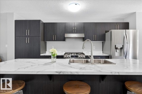 Kitchen featuring a large island with a double basin sink and a high-arc faucet, dark wood-finish cabinetry, stainless steel appliances, and light-toned countertops - 3043 Keswick Way Sw, Edmonton, AB - Indoor Photo Showing Kitchen With Stainless Steel Kitchen With Double Sink With Upgraded Kitchen