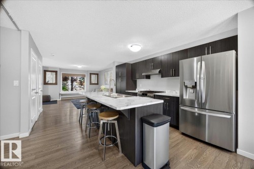 Open-concept kitchen featuring an oversized island with seating, dark wood-finish cabinetry, stainless steel appliances, and wood-finish flooring - 3043 Keswick Way Sw, Edmonton, AB - Indoor Photo Showing Kitchen With Stainless Steel Kitchen With Upgraded Kitchen