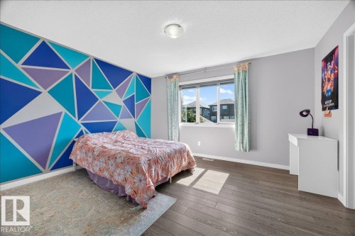 Spacious room featuring wood-finish flooring, a geometrically patterned accent wall, and a large window with white trim - 3043 Keswick Way Sw, Edmonton, AB - Indoor Photo Showing Bedroom