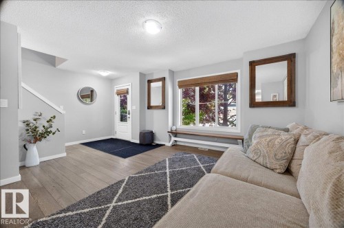 Bright living area featuring light gray walls, wood-finish flooring, a large window with natural light, a built-in window bench, and a white entry door - 3043 Keswick Way Sw, Edmonton, AB - Indoor