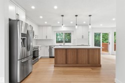 Modern kitchen featuring a wood-panel island with a light-toned countertop, stainless steel appliances, white shaker cabinetry with black hardware, and light wood-finish flooring - 