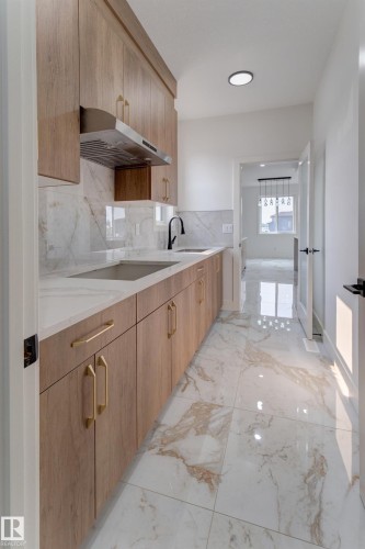 Contemporary galley kitchen featuring wood-finish cabinetry with golden hardware, a large stainless steel sink with a matte black faucet, and stone-look slab countertops - 4723 17 Avenue Sw, Edmonton, AB - Indoor