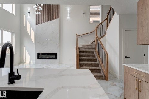 Spacious interior featuring a contemporary fireplace with a decorative wood accent wall, a grand staircase with wood railings and black spindles, integrated stair lighting, large windows, and sleek white flooring - 4723 17 Avenue Sw, Edmonton, AB - Indoor Photo Showing Other Room With Fireplace