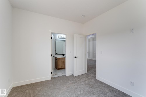 Neutral gray carpet flooring complements clean white walls in this room, featuring a doorway opening to a bathroom with a wood-finish vanity, a rectangular mirror with a matte black frame, and a three-light fixture - 4723 17 Avenue Sw, Edmonton, AB - Indoor Photo Showing Other Room