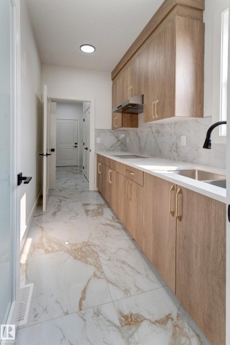 Contemporary galley kitchen featuring wood-finish cabinetry with gold-tone hardware, white countertops, and a white and gold patterned tile backsplash - 4723 17 Avenue Sw, Edmonton, AB - Indoor Photo Showing Kitchen
