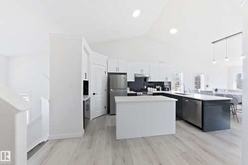 Modern kitchen featuring light wood-finish flooring, vaulted ceilings, white cabinetry, a dark stone-look backsplash, and a large central island with a built-in sink and stainless steel appliances - 3799 21 Street Nw, Edmonton, AB - Indoor Photo Showing Kitchen