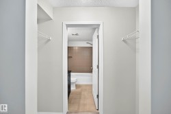 Hallway with light gray walls and white wire shelving, offering entry to a bathroom featuring a white tub, brown tiled shower surround, and tan tile flooring - 