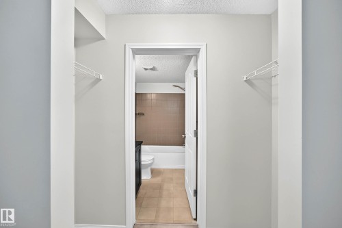 Hallway with light gray walls and white wire shelving, offering entry to a bathroom featuring a white tub, brown tiled shower surround, and tan tile flooring - #404 12025 22 Av Sw, Edmonton, AB - Indoor
