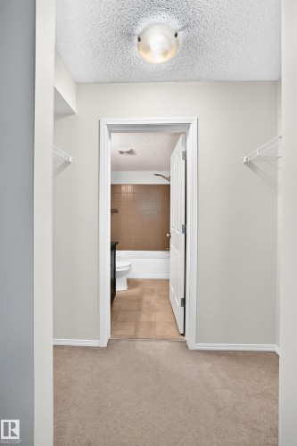 Hallway with neutral tone carpeting and white trim leading to a bathroom featuring a white bathtub, ceramic tile shower surround, and tile flooring - #404 12025 22 Av Sw, Edmonton, AB - Indoor